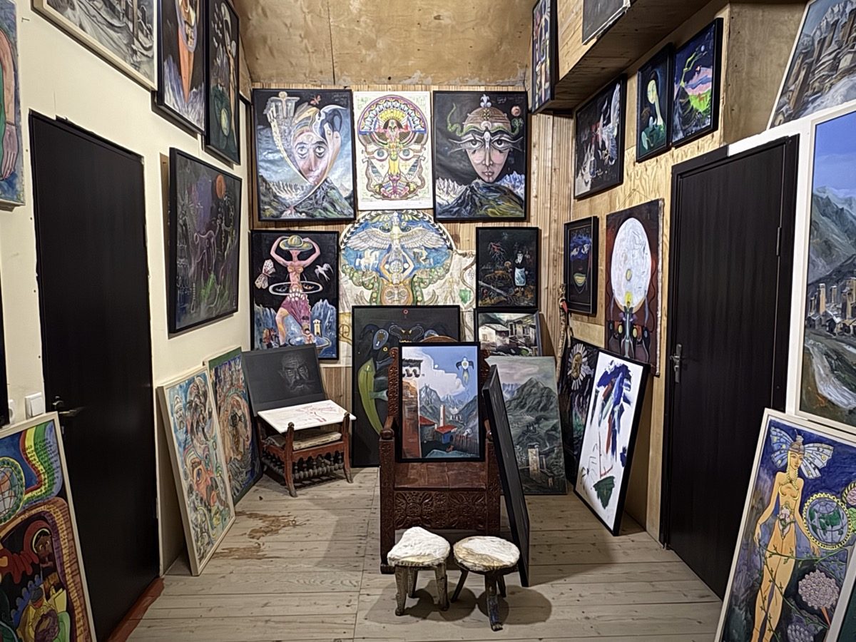 A small room with bare wooden walls and floor, hung from floor to ceiling with framed paintings: faces, figures, suns and mountains. More canvases stand leaning against the walls, beside a carved wooden chair and two carved stools.
