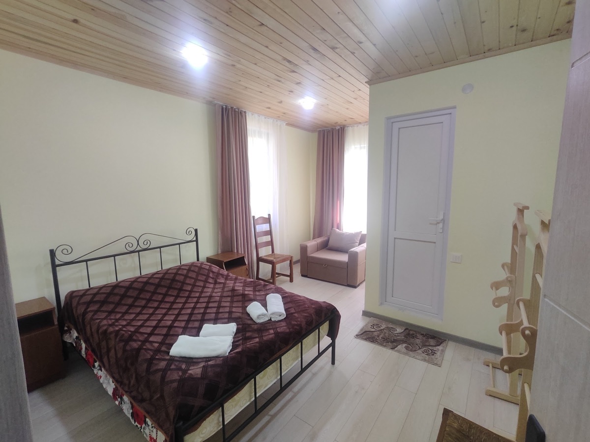 An upstairs guest room with a double bed, wooden ceiling, two windows and a chair beside the bed.