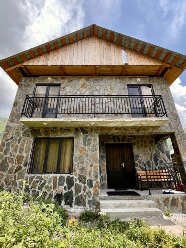 The guesthouse: a two-storey building of grey and ochre stone under a wide wooden gable, with a balcony running the width of the upper floor and a bench beside the front door.